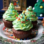 3 Christmas tree cupcakes on a festive plate with partially-visible Christmas decorations in the background.