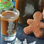 A glass shot with gingerbread syrup with a bottle of syrup and a gingerbread man cookie behind it and Christmas decorations around.