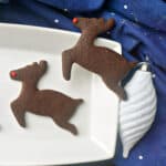 2 reindeer-shaped cookies on a white plate with a white Christmas bauble and a blur tea towel around the plate.