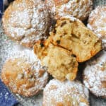 A plate with 2 halves of a muffin on top of other 6 muffins dusted with icing sugar and a blue tea towel around the plate.