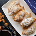 A white rectangle plate with almond croissants, whole almonds and a cup of coffee with a saucer next to the plate.