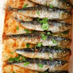 A white dish with baked sardines garnished with chopped parsley.