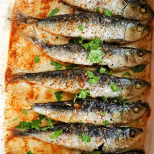 A white dish with baked sardines garnished with chopped parsley.