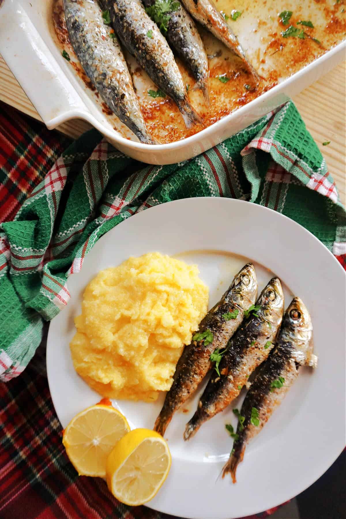 A white plate with 3 baked sardines, polenta and 2 slices of lemon, a dish with more sardines and a green kithcen towel around them.