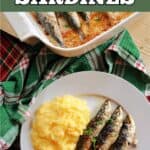 A white plate with 3 bakes sardines, polenta and 2 slices of lemon, a green tea towel and a partially visible white dish with more sardines.