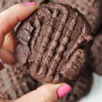 A hand holding a chocolate cookie with other blurred cookies in the background.