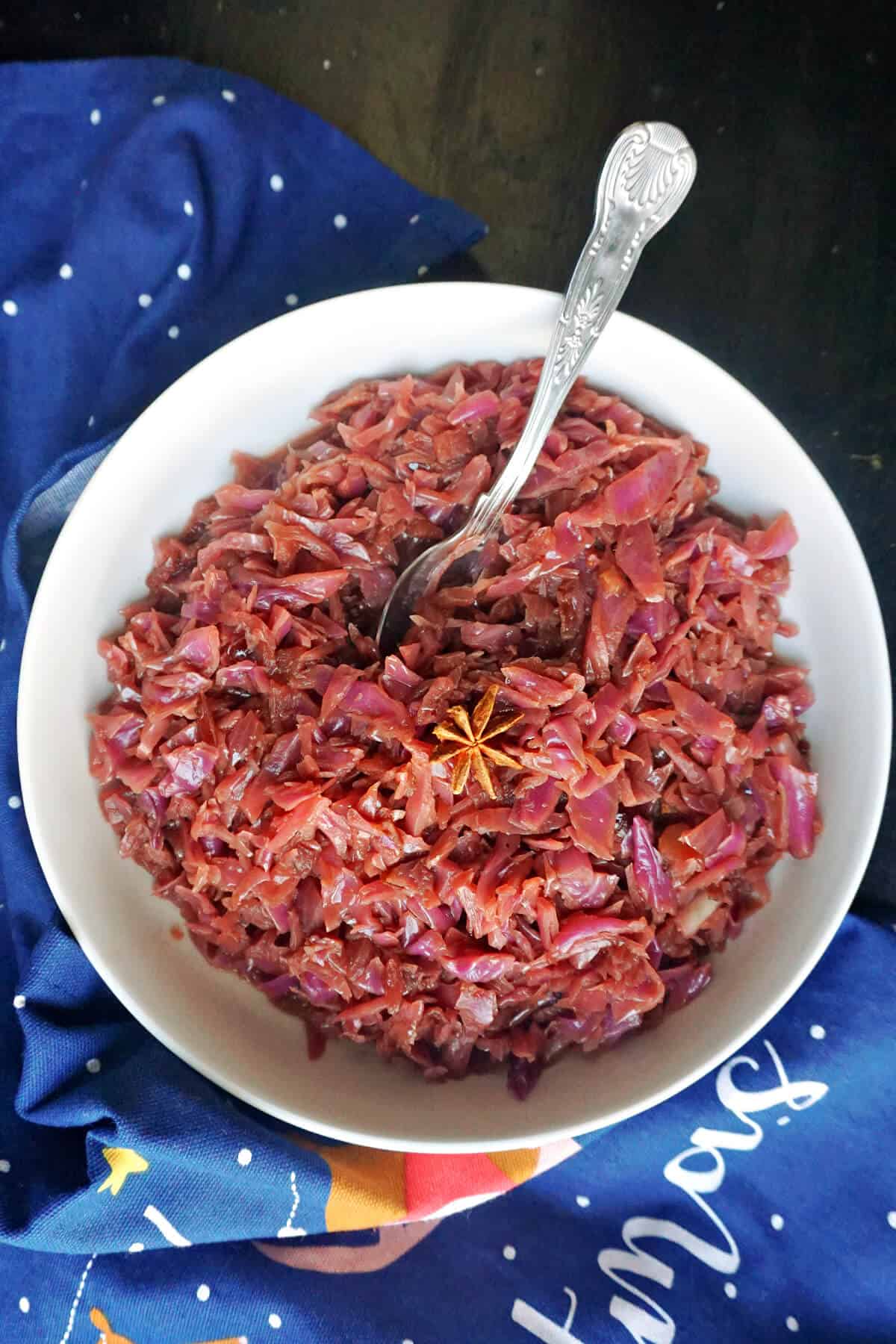 A white bowl with braised red cabbage topped with a star anise and a spoon in the cabbage. Around the bowl there is a blue kitchen towel.