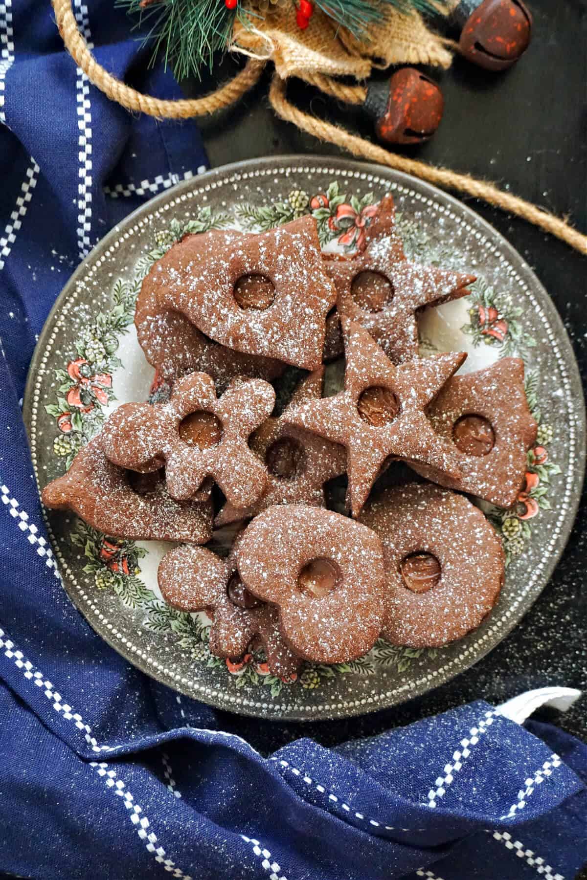A festive plate with nutella-stuffed chocolate cookies dusted with icing sugar, a blue and white kitchen towel around the plate and a Christmas decoration around.