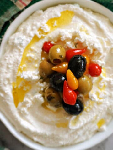 A white bowl with a cottage cheese dip topped with olives, yellow and red peppers and olive oil. A green tea towel around it.