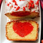 A slice of a cake with a red heart inside, the rest of the sliced cake decorated with icing and red crumbs on a white rectangle plate.
