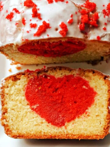 A slice of a cake with a red heart inside, the rest of the sliced cake decorated with icing and red crumbs on a white rectangle plate.