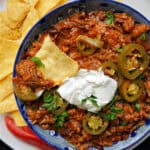 A blue plate with chili topped with sour cream, sliced jalapenos and a tortilla chip and more chips on the side.