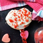 A heart-shaped cake pop with red crumbs on top, smal pink and red hearts and a pink tea towel around.
