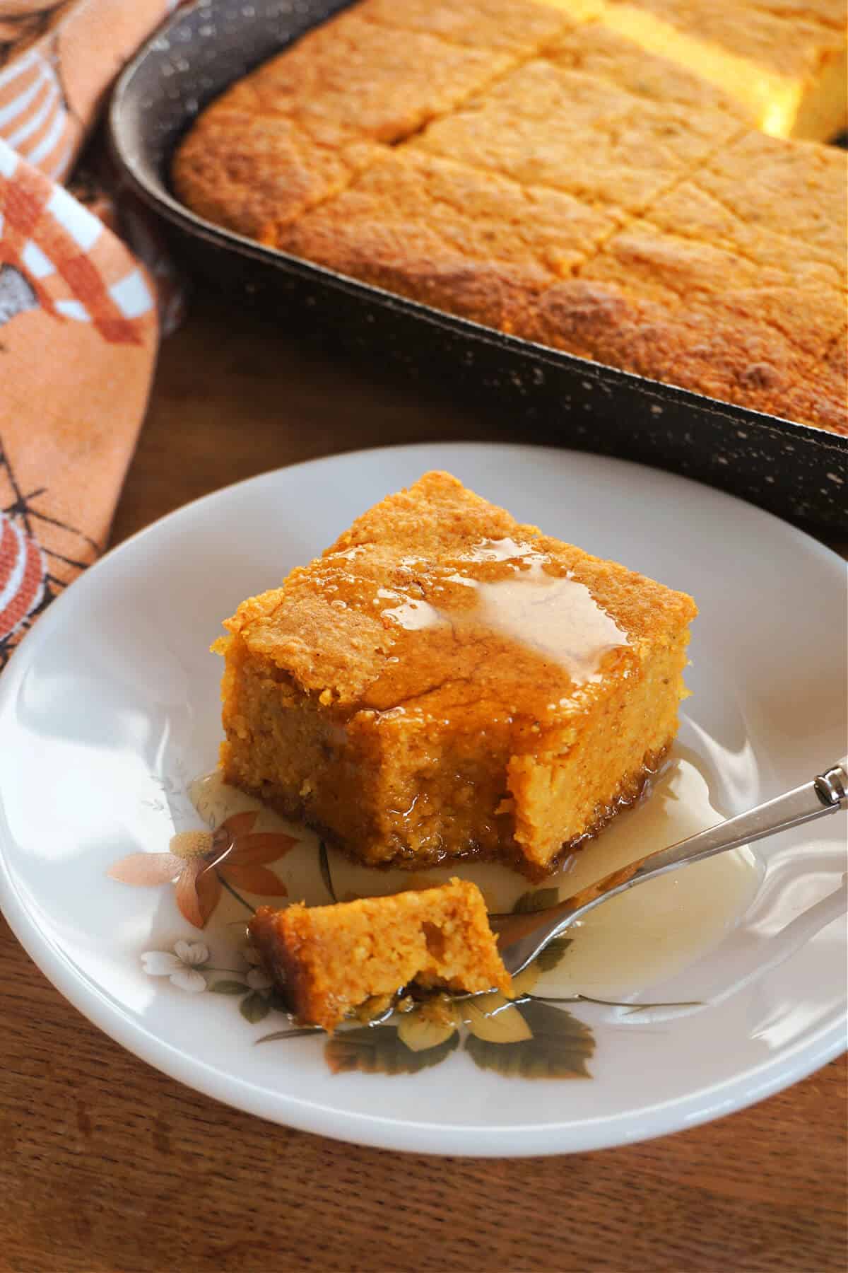 A dessert plate with a slice of pumpkin cornbread drizzled with maple syrup and a fork with a bite of cornbread on it. In the background there is a pan with cornbread and an orange patterned tea towel.