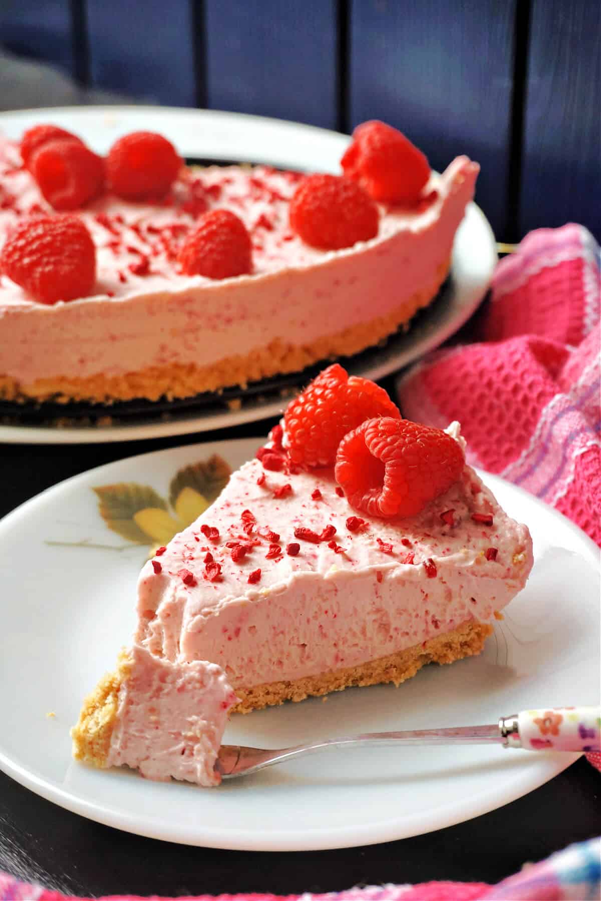 A white plate with a slice of raspberry cheesecake topped with 2 raspberries and freeze-dried raspberries, a dessert fork with a chunk of cheesecake and a plate with the remaining cheesecake in the background.