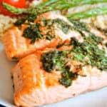 A plate with 2 cooked salmon fillets topped with herbs, and partially visible asparagus and tomatoes on a bed of couscous.