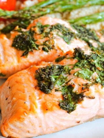 A plate with 2 cooked salmon fillets topped with herbs, and partially visible asparagus and tomatoes on a bed of couscous.