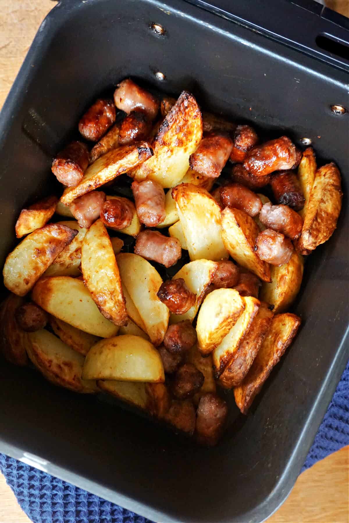 An air fryer drawer with cooked potato wedges and sausage chunks.