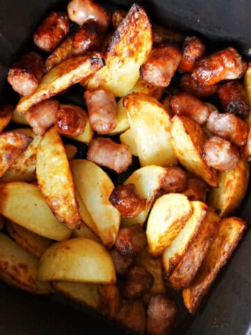 An air fryer drawer with cooked potatoe wedges and sausage cut into chunks.