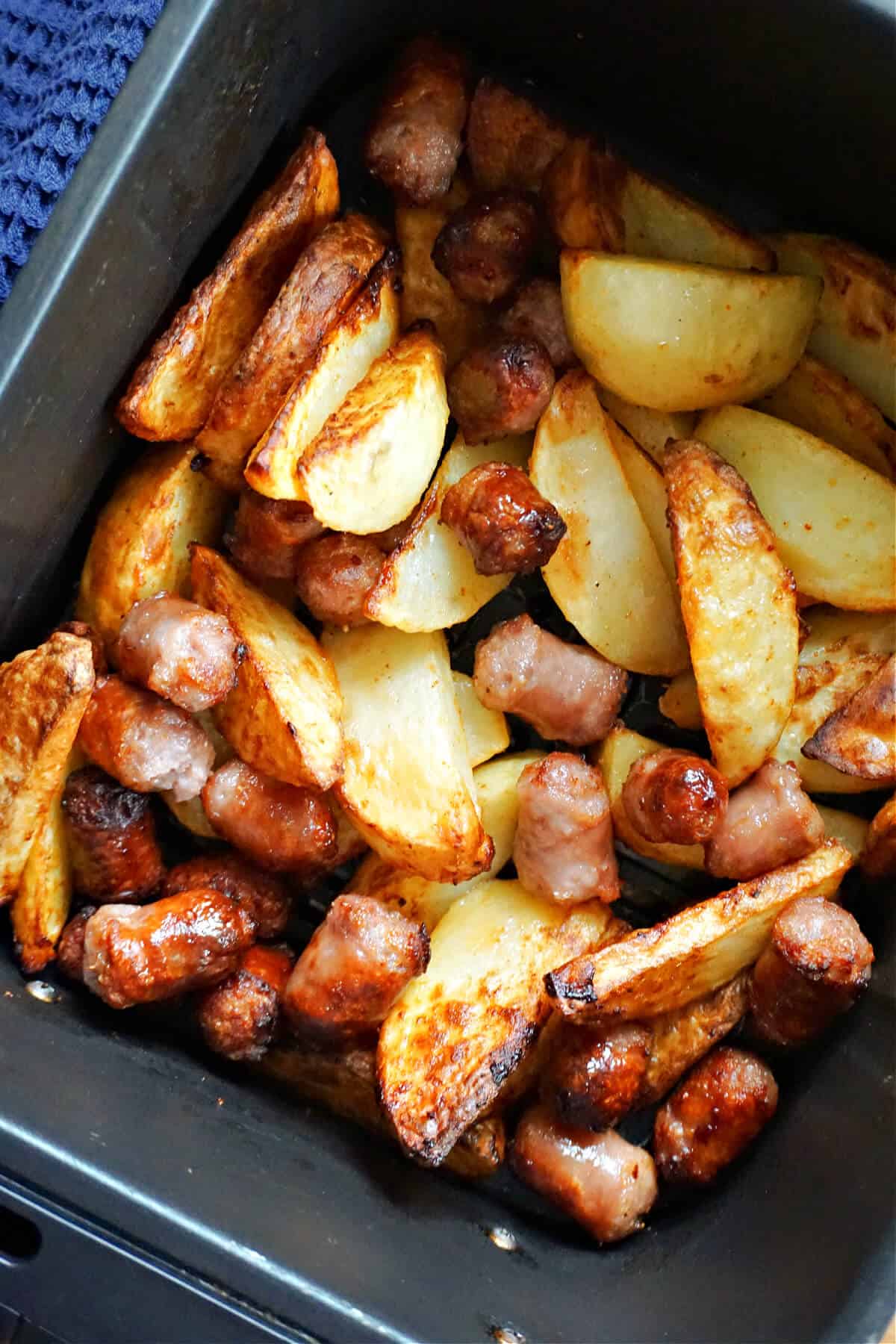 An air fryer drawer with potatoes wedges and chopped sausages.
