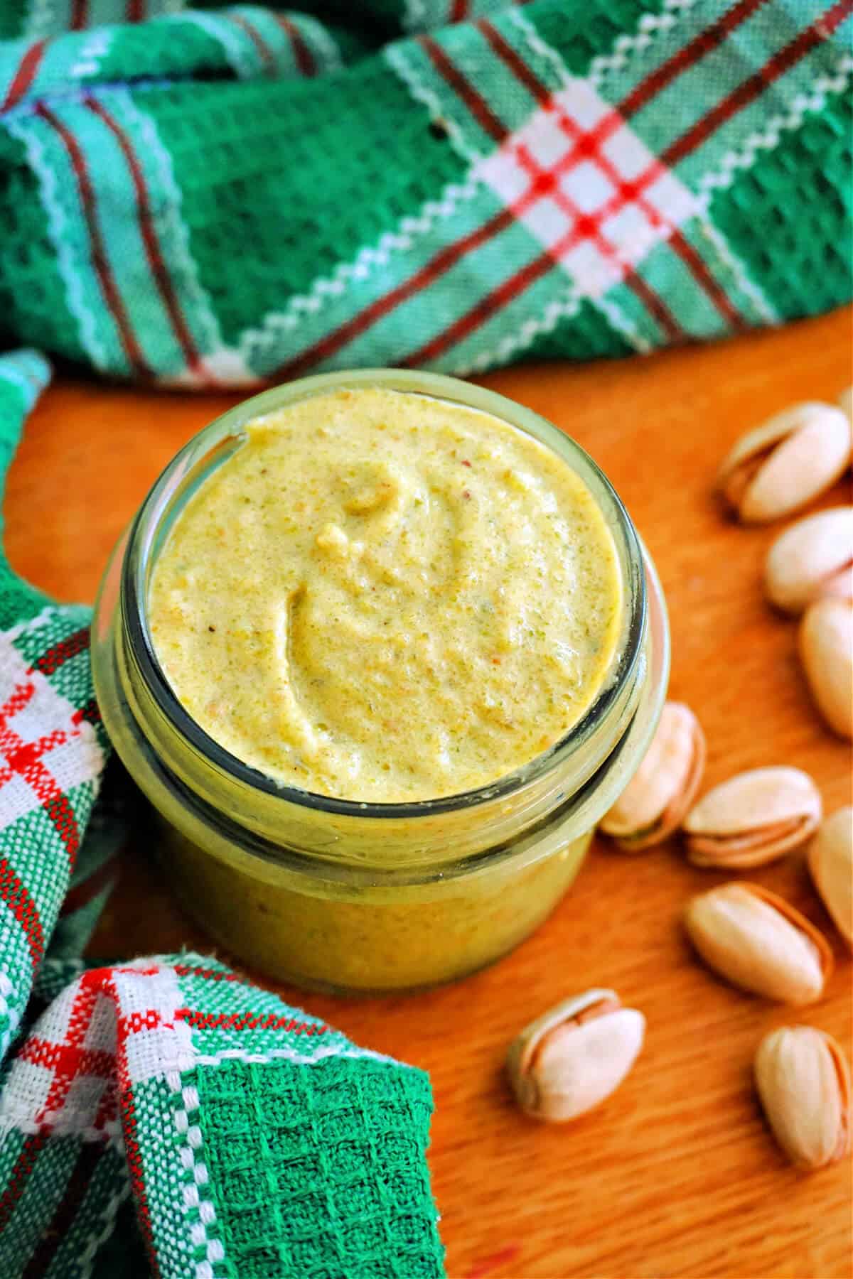 A jar with pistachio cream with pistachios around it and a green patterned tea towel.