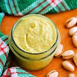 A jar with pistachio cream, shell-on pistachios and a green-patterned tea towel around the jar.