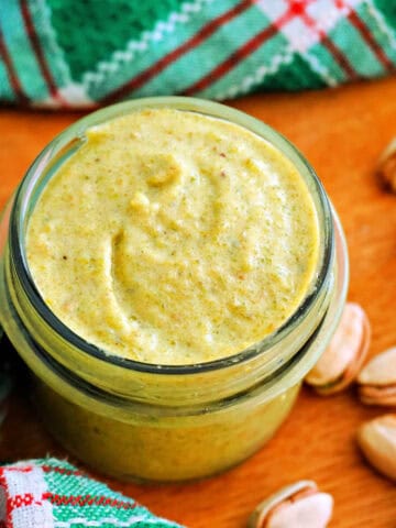A jar with pistachio cream, shell-on pistachios and a green-patterned tea towel around the jar.