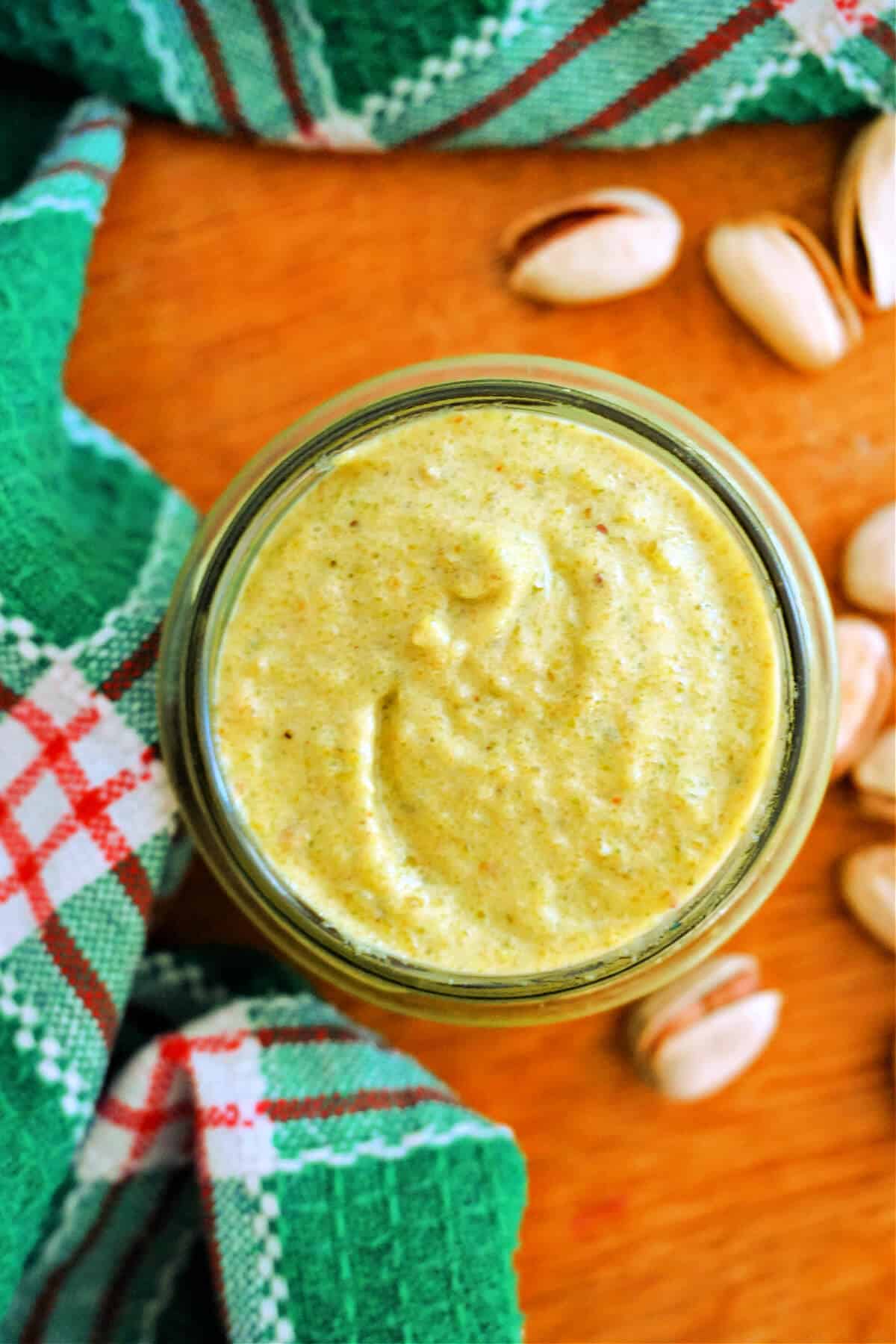 Overhead of a jar with pistachio cream, pistachios around the jar and a green-patterned tea towel.