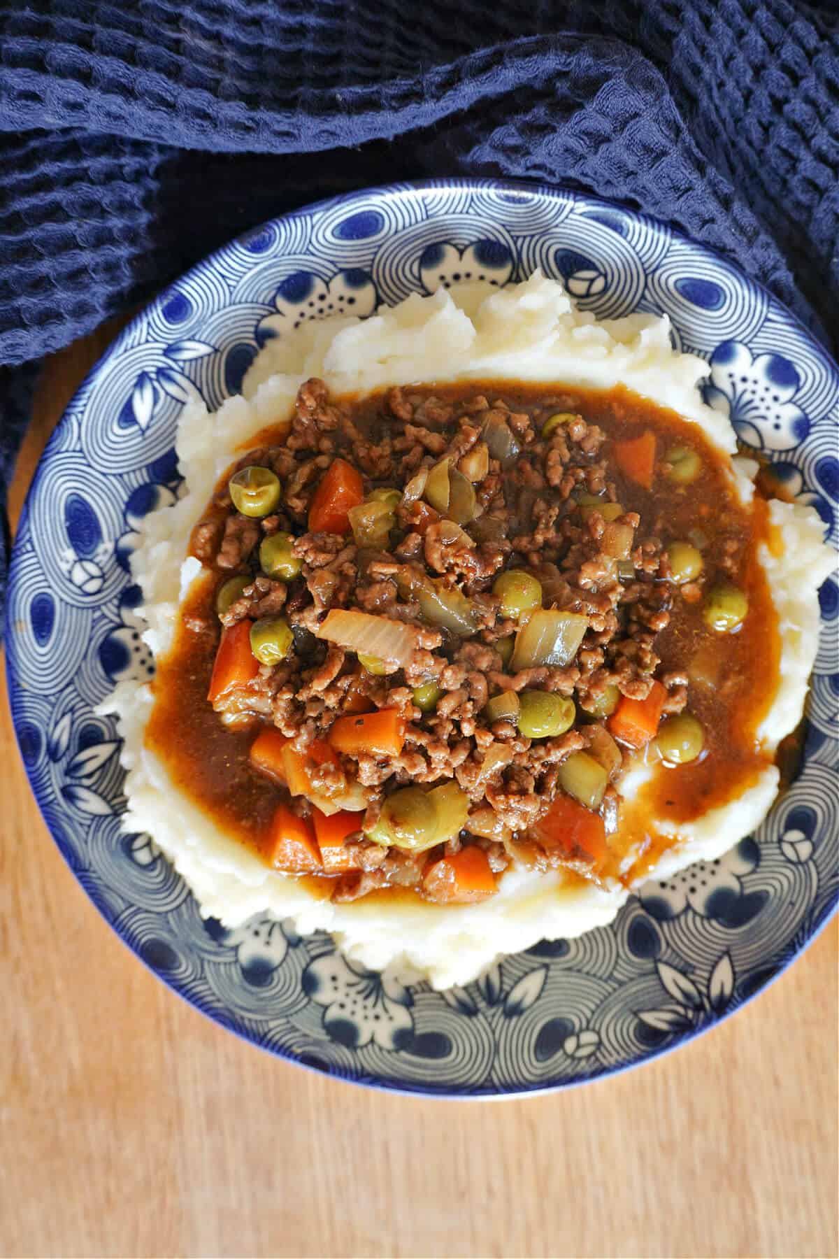 Overhead of a blue patterned plate with mince and gravy with veggies over a bed of mash.