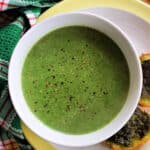 A white bowl with creamy nettle soup on a white plate, 2 crostini on the plate and a green tea towel around the plate.