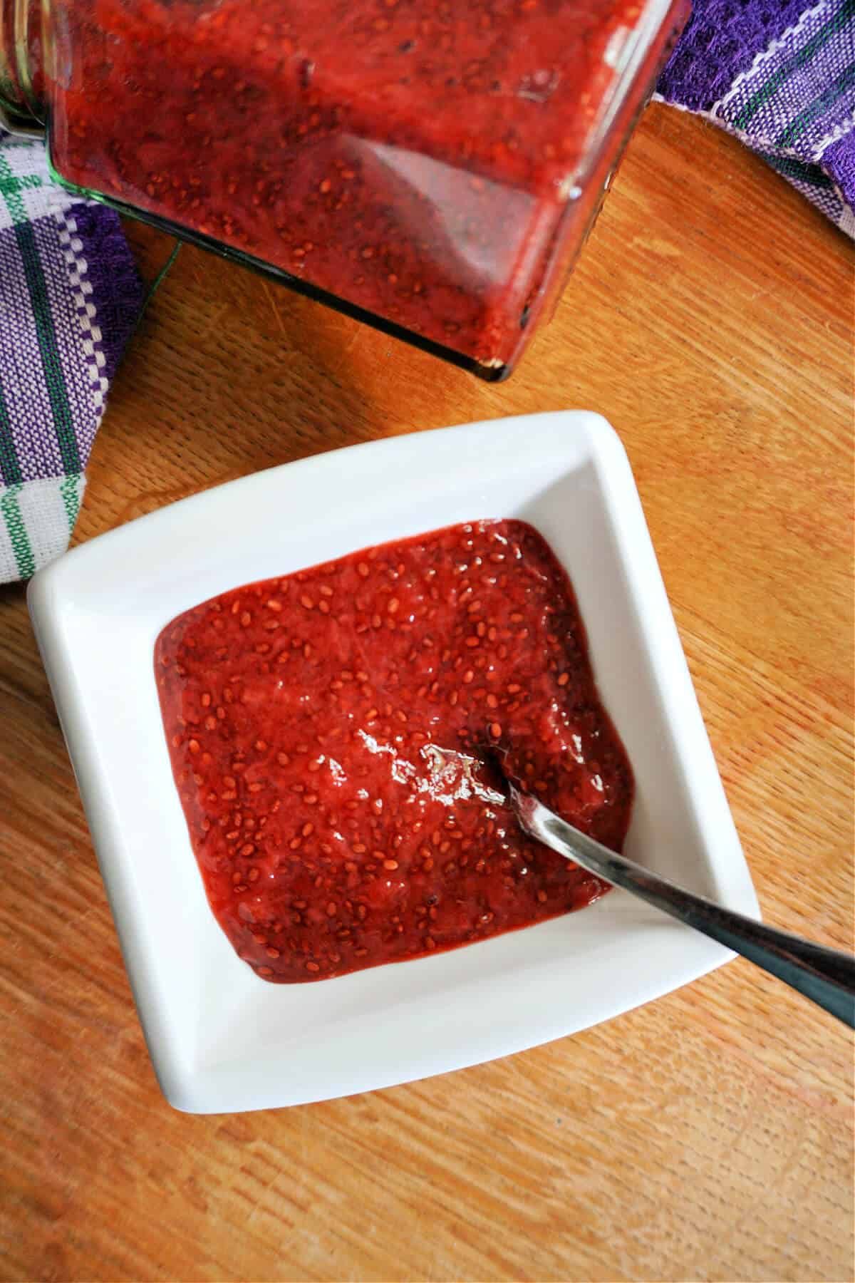 A white ramekin with strawberry jam, a jar with jam and a purple-patterned tea towel around it.