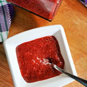 A white ramekin with jam and teaspoon in it, a jar of jam and a partially-visible tea towel next to it.