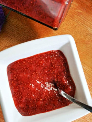 A white ramekin with jam and teaspoon in it, a jar of jam and a partially-visible tea towel next to it.