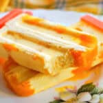 A plate with a mango popsicle with a bite off it on top of another whole popsicle.