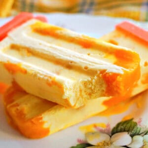 A plate with a mango popsicle with a bite off it on top of another whole popsicle.