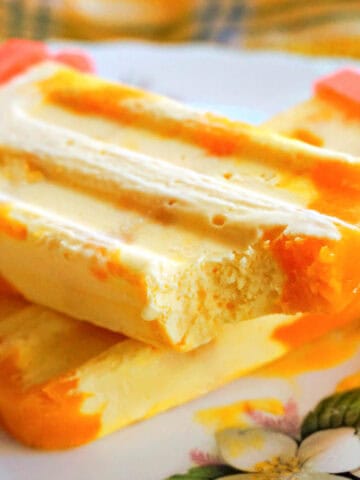 A plate with a mango popsicle with a bite off it on top of another whole popsicle.