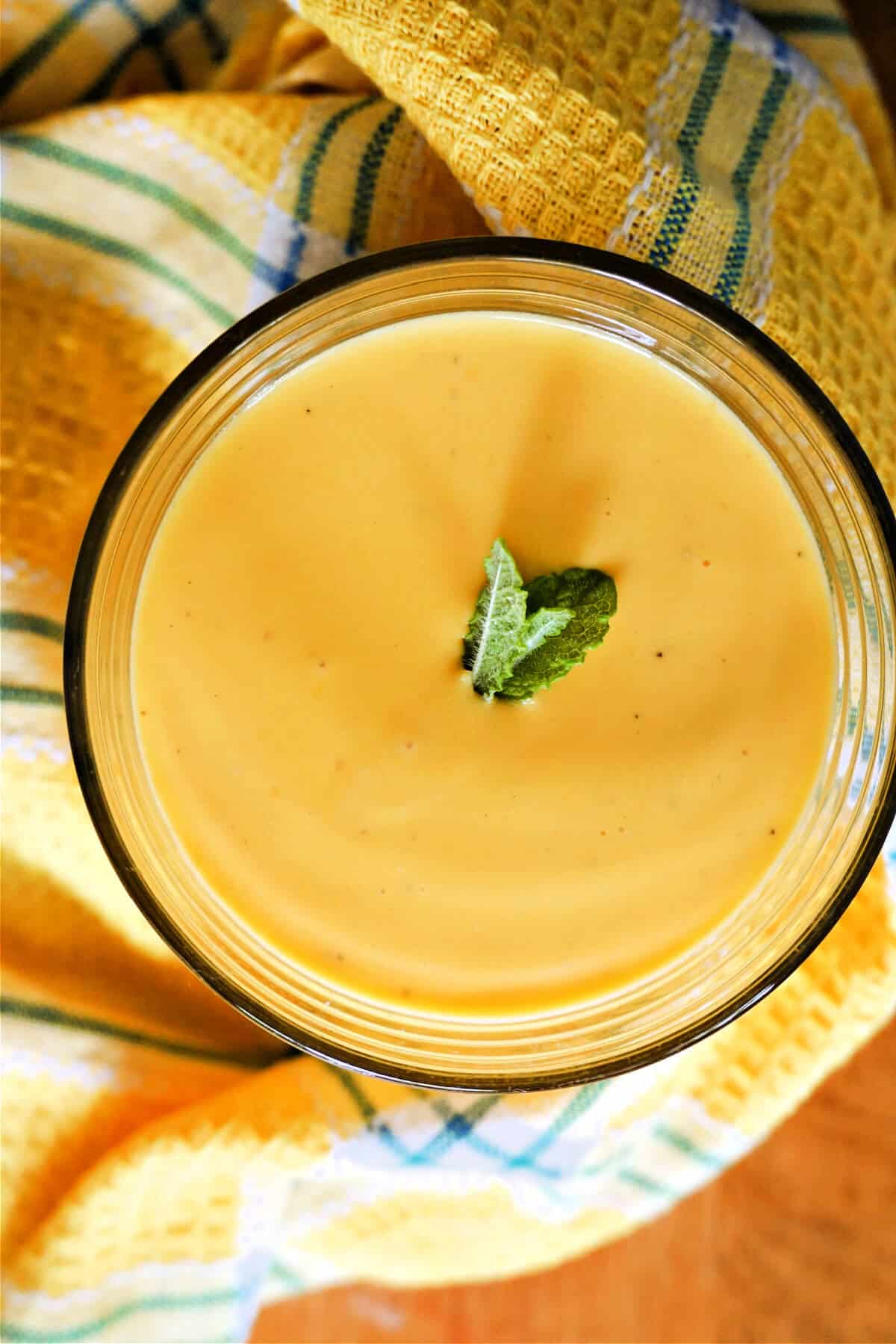 Overhead of a glass with mango lassi garnished with minit leaves and a yellow-pattered tea towel around it.