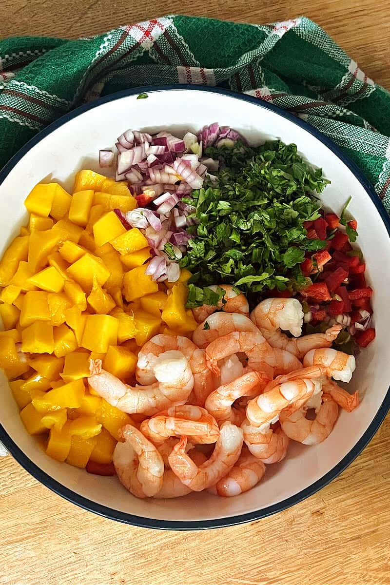 A bowl with choped mango cubes, chopped red onion, cooked prawn, chopped parsley and chopped red peppers. A green tea towel around it.