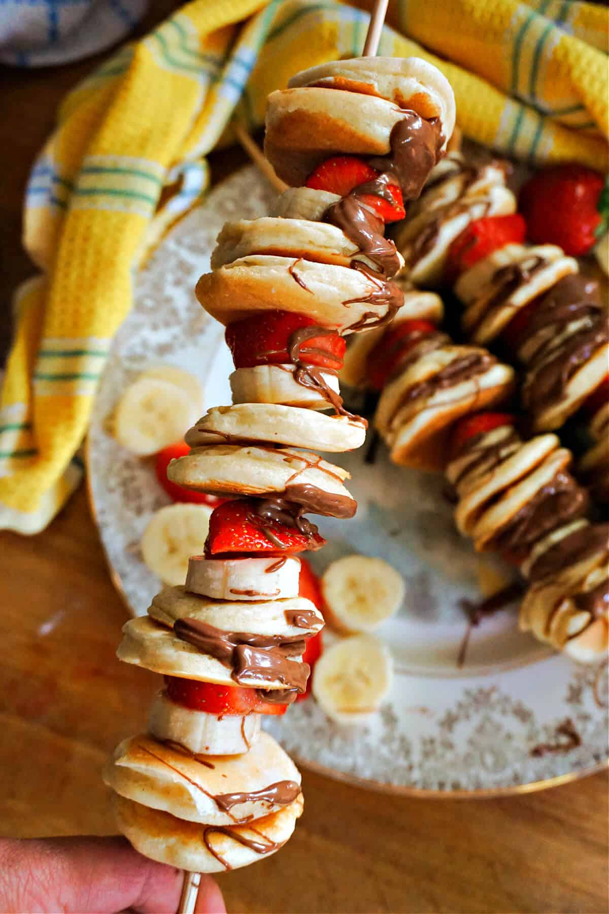 A pancake skewer with mini pancakes, banana and strawberry slices and 2 othe skewers in the background on a plate. A yellow-patterned tea towel is around the plate.