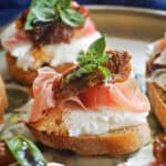 A slice of bruschetta with mozzarella, prosciutto, sun-dried tomato and basil and other bruschettas partially-visible on the plate.