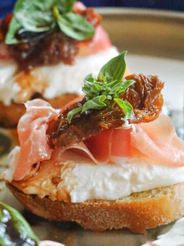 A slice of bruschetta with mozzarella, prosciutto, sun-dried tomato and basil and other bruschettas partially-visible on the plate.