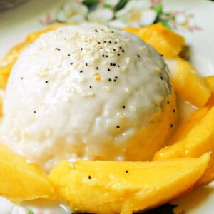 A creamy rice dessert topped with sesame seeds, surrounded by fresh mango slices on a decorative plate.