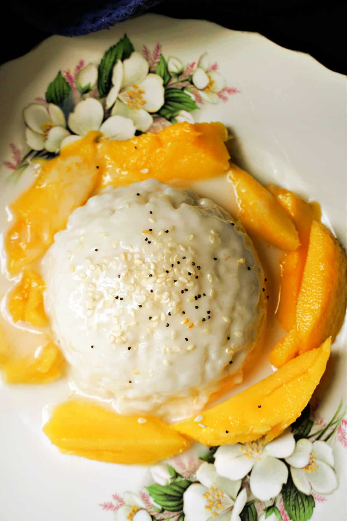 A white flowery plate with a home-shaped portion of rice garnished with seasame and poppy seeds and mango slices around it.