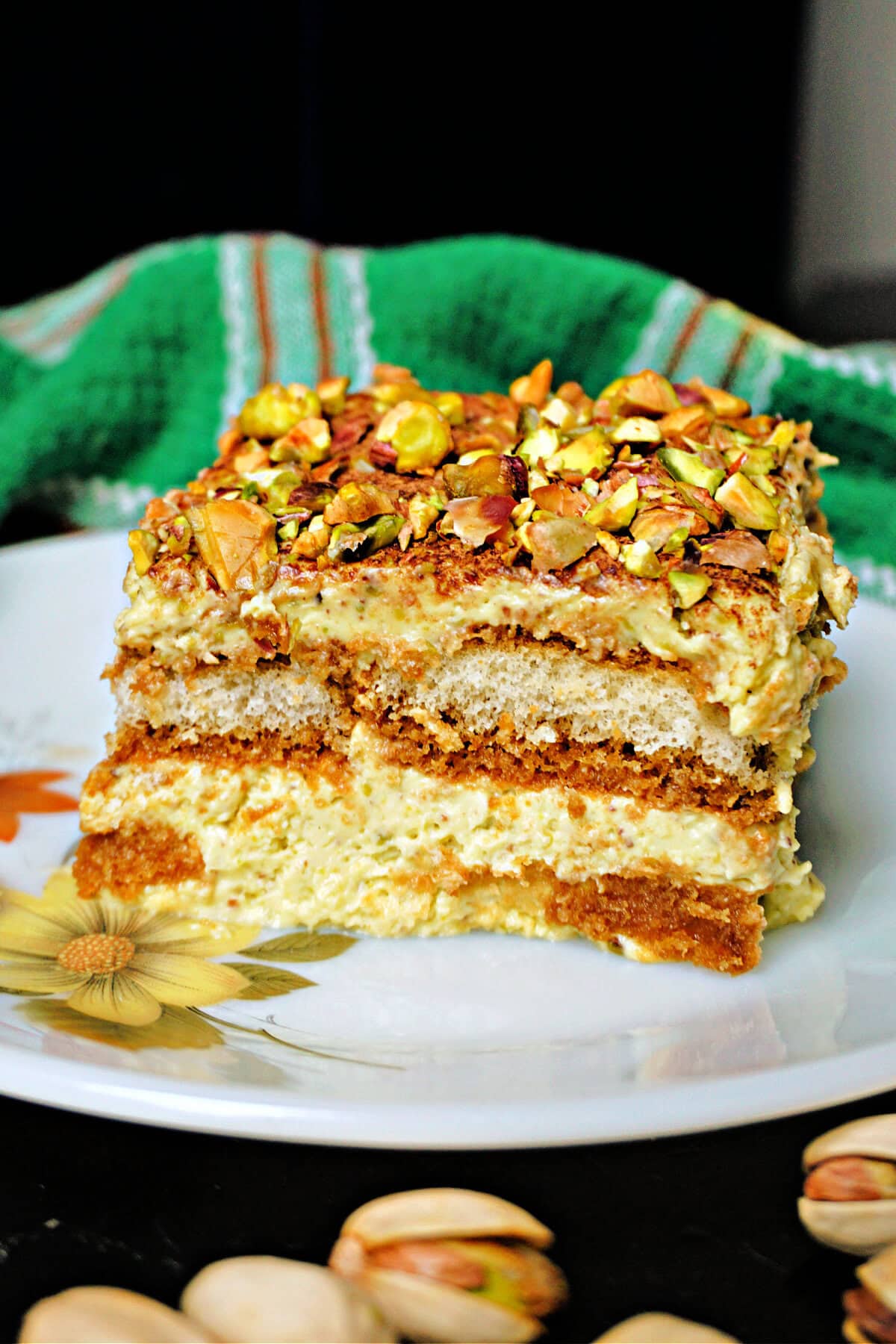 A slice of pistachio tiramisu on a white plate with a green tea towel in the background.