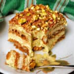 A slice of pistachio tiramisu on a white plate with a dessert fork resting on it and a green tea towel in the background.