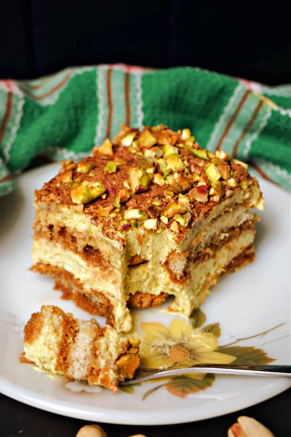 A white flowery plate with a slice of pistachio tiramisu and a silver fork with a piece of cake resting on it.