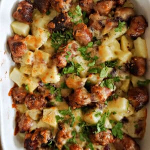A white casserole with sausages and potatoes topped with melted cheese and chopped parsley.