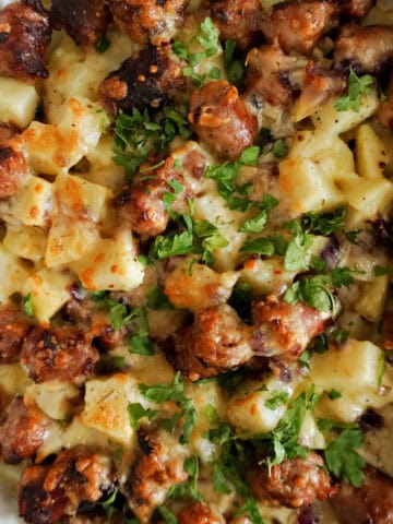 A white casserole with sausages and potatoes topped with melted cheese and chopped parsley.