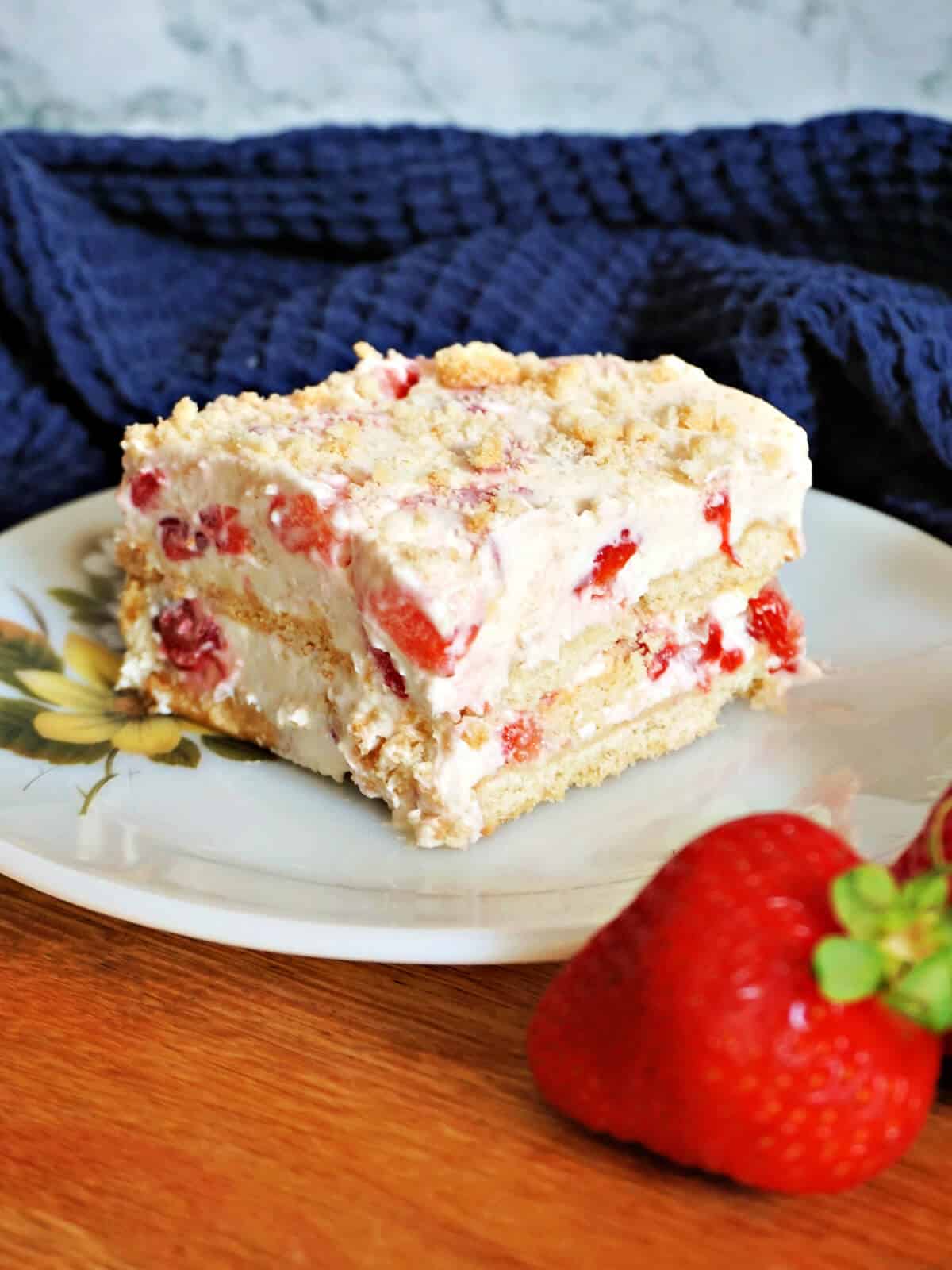 A white plate with a slice of strawberry cake and a strawberry by the plate. A navy blue tea towel in the background.