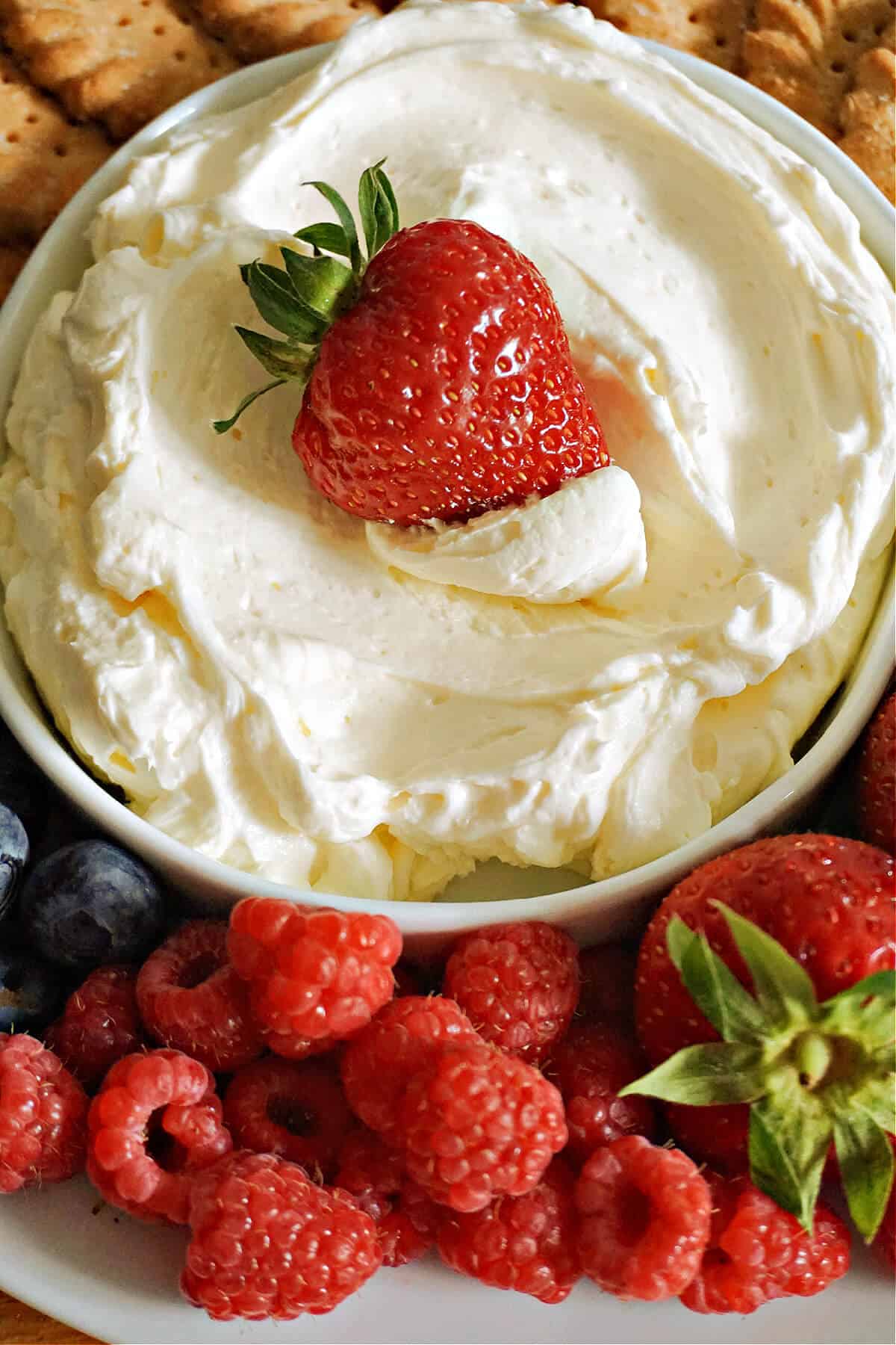 A white bowl with cheesecake dip and a strawberry dipped into it, and other berries around the bowl.